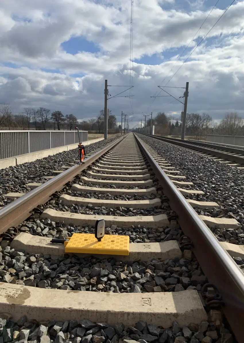 Gerade verlaufendes Bahngleis mit Oberleitungen, Schotterbett und gelbem Messgerät auf einer Betonschwelle. Erprobung SNS
