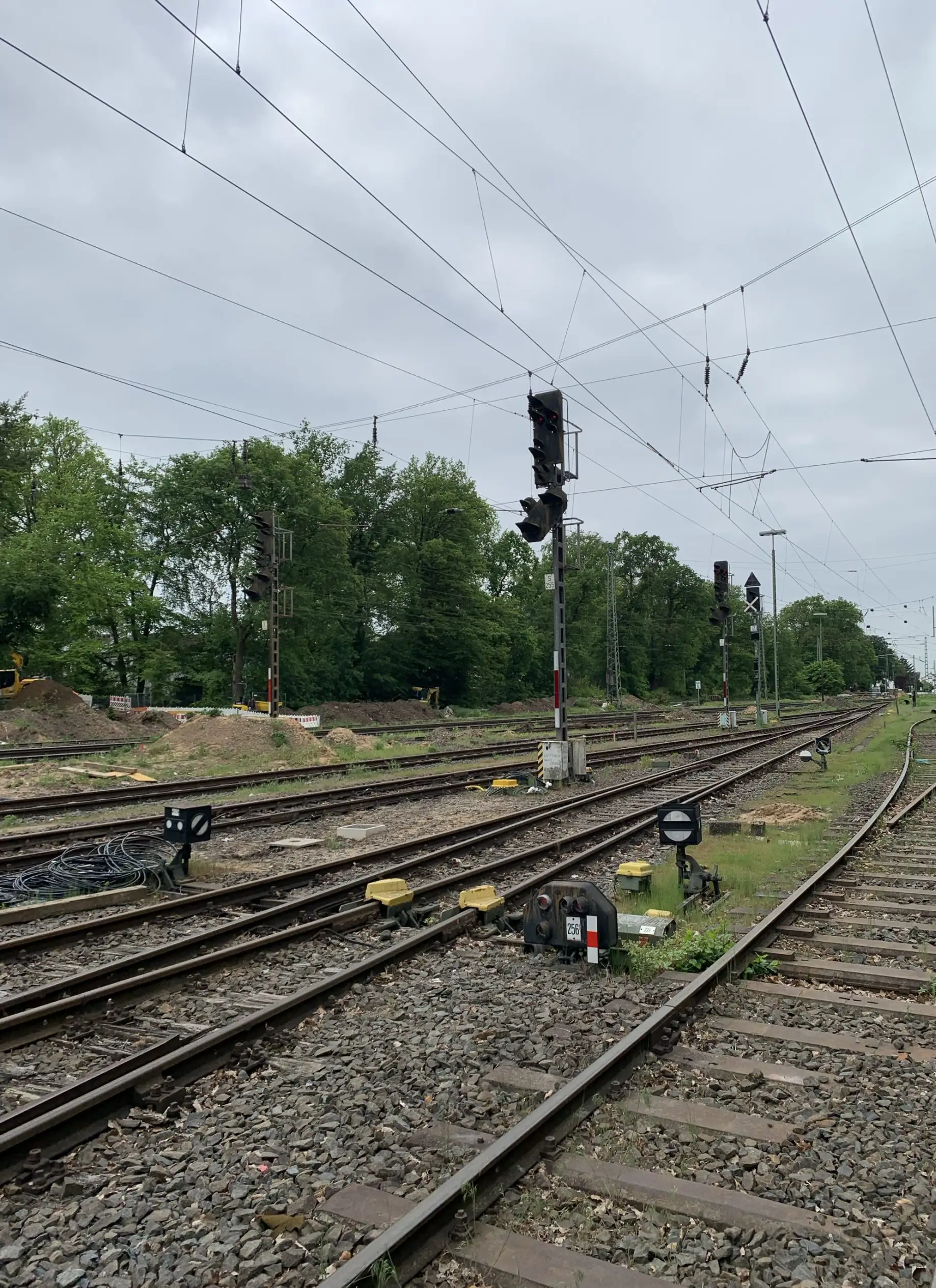 Gleisfeld mit Signalen, Oberleitungen, Bäumen und Bauausrüstung am Rand.