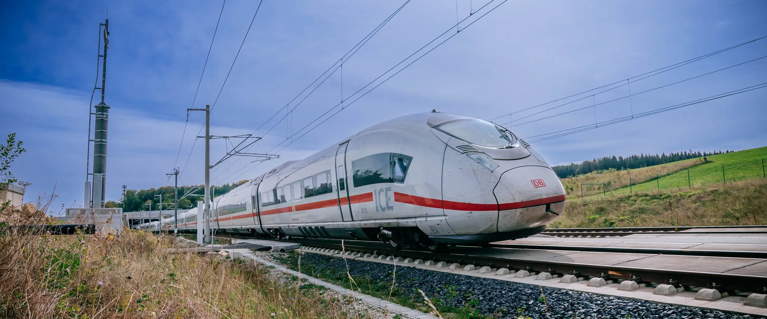 Weißer ICE-Zug mit rotem Streifen auf Gleisen in ländlicher Umgebung mit Oberleitungen.